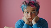 Young person with colorful dyed hair in blue sweater looking thoughtful with hands on face against pink background for youth lifestyle concepts.