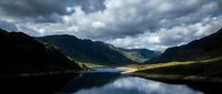Dramatic mountain valley landscape with serene lake reflecting stormy clouds and sunlit peaks, suitable for travel, nature, and environmental design projects.