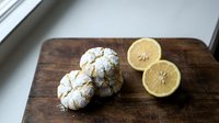 Freshly baked lemon crinkle cookies with powdered sugar dusting on rustic wooden cutting board alongside halved lemon for baking and cooking content.