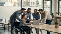 Diverse business team collaborating around laptop in modern office with wheelchair-accessible workspace promoting workplace inclusion and teamwork.