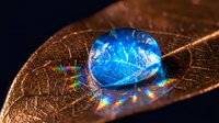 Blue water droplet with rainbow reflections resting on golden autumn leaf surface creating prismatic light effects and natural macro photography composition.
