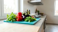 Fresh vegetables including tomatoes, eggplant and herbs arranged on turquoise cutting board in modern kitchen interior for healthy cooking preparation.