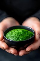 Hands holding black ceramic bowl with vibrant green matcha tea powder against dark background, dramatic lighting highlights tea ceremony and wellness concept.