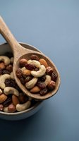 Mixed nuts and dried fruits in wooden spoon and bowl on blue background. Healthy snacking, trail mix with almonds, cashews, raisins for nutrition.