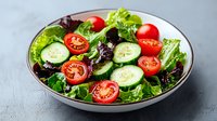 Fresh garden salad with sliced cucumbers, cherry tomatoes and mixed lettuce leaves in gray ceramic bowl on light background, healthy eating concept.