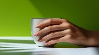 Female hand with manicured nails gently holding white ceramic cup on white surface against vibrant green background with dramatic lighting and shadow pattern.