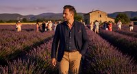 Middle-aged Caucasian man in casual blazer walking through purple lavender field at sunset with tourists and rustic building in background.