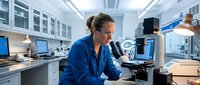 Female scientist in blue lab coat working with microscope in modern research laboratory with computers and equipment for medical analysis.