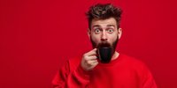 Surprised young Caucasian man with beard drinking coffee from black mug against vibrant red background, wearing matching red sweater.