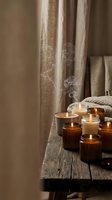 Lit candles in glass containers arranged on wooden surface with draped linen curtains and woven textile, creating warm ambient lighting for home decor, relaxation, wellness, and interior design projects.