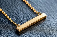 Golden horizontal bar pendant necklace on textured dark slate background, minimalist jewelry design for fashion and accessory marketing.