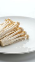 Enoki mushrooms arranged on white plate, fresh raw fungi with delicate stems and small caps, suitable for culinary and food photography.