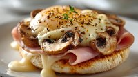 Gourmet breakfast sandwich with poached egg, melted cheese, ham, and sautéed mushrooms on toasted English muffin, garnished with herbs and spices.