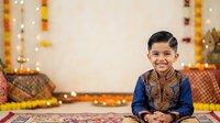 Young South Asian boy, approximately 6-8 years old, wearing traditional blue kurta with gold embroidery, smiling at camera in festive setting with marigold garlands and decorative lights for Diwali or wedding celebration.