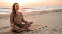 Young Caucasian woman meditating in lotus pose on sandy beach at sunset, wearing linen clothes, eyes closed, peaceful mindfulness practice.