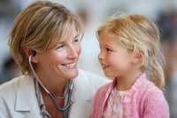 Caring female doctor with stethoscope smiling at young blonde girl patient during medical examination, creating warm and trusting healthcare environment.