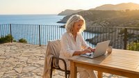 Mature woman with white hair working on laptop at wooden table on coastal terrace overlooking Mediterranean sea at sunset, remote work and travel lifestyle.
