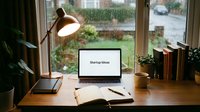 Laptop on a wooden desk displaying startup ideas text, with a lamp, open notebook, books, and plants by a rainy window, cozy home office.