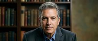 Mature man with gray hair and light skin tone wearing dark blazer and blue shirt, seated in front of home library bookshelf, professional portrait for business, education, or corporate use.
