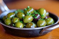 Fresh marinated green olives with herbs and spices served in rustic cast iron pan on wooden table, close-up view showing glossy texture and garnish.
