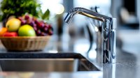 Modern chrome faucet in luxury kitchen interior with fresh fruits in wicker basket on counter, selective focus on tap with blurred background.