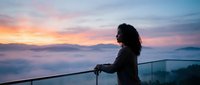 Woman with long dark curly hair standing at railing overlooking misty mountain valley at sunrise, wearing dark jacket, contemplative pose for travel and lifestyle content.