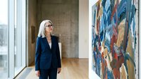 Professional mature Caucasian businesswoman in navy blazer admiring contemporary abstract artwork in modern gallery space with concrete walls and wooden floors.