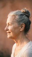 Senior woman with gray hair styled in a bun, smiling in profile against warm neutral background, wearing light colored clothing for portrait and lifestyle use.