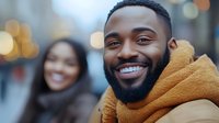Happy young African American man with beard smiling at camera wearing orange hoodie on city street with blurred woman in background during daytime.