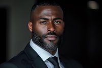 Professional African American male executive in black suit and tie with well-groomed beard looking confidently at camera against dark background, portraying leadership and success.