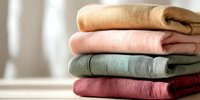 Stack of folded soft linen towels in earthy tones of beige, pink, sage green, and burgundy on wooden surface, creating cozy bathroom or spa atmosphere.