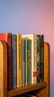 Row of classic literature books arranged on wooden shelf against gradient pink and blue background, featuring vintage spines with gold lettering for reading and library design.