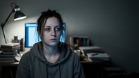 Young woman with messy hair wearing headphones and hoodie in dimly lit room with desk lamp and books, late night study or gaming session atmosphere.