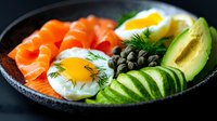 Fresh salmon with sunny side up eggs, sliced avocado, capers, cucumber and dill garnish served in dark ceramic bowl for healthy breakfast or brunch.