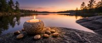 Carved wooden candle holder with burning flame surrounded by smooth stones on rocky shore at peaceful lake sunset with pine trees and purple sky reflection.