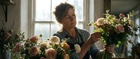 Mature woman florist arranging fresh roses and greenery in vases by bright window, creating floral arrangements for events, weddings, or home decor.