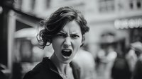 Young Caucasian woman shouting in anger on city street, black and white portrait capturing intense emotional expression against blurred urban background.