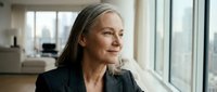 Confident mature businesswoman with gray hair looking out office window, professional executive in corporate environment planning future success.