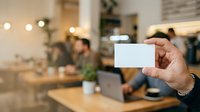 Businessman holding blank white business card in modern office workspace with colleagues working in blurred background, networking and professional communication concept.