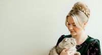 Young woman with blonde hair styled in an updo, fair complexion, holding and petting a gray tabby cat. She wears a dark green top and smiles warmly while looking at the animal. Isolated on white background for pet care, veterinary, and lifestyle design use.