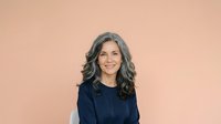 Middle-aged woman with long wavy gray hair and warm smile wearing dark navy blue top against soft peachy beige background. Portrait suitable for business, healthcare, corporate websites, and professional profiles.