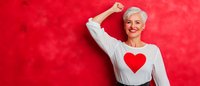 Energetic senior woman with short gray hair wearing white sweater with red heart design celebrates with raised fist against vibrant red background.
