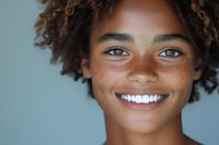 Young African American woman with natural curly hair and freckles showing bright white smile against blue background, close up portrait expressing joy and confidence.