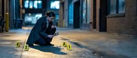 Forensic investigator examining evidence at urban crime scene with numbered markers during nighttime investigation in city street.