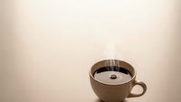 Brown ceramic coffee cup with steaming hot black coffee on neutral beige background with copy space for morning beverage and cafe concepts.