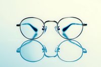 Round metal frame glasses with reflection on light blue surface, minimalist composition showing eyewear design and optical properties in studio lighting.