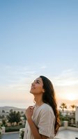 Young woman with long dark hair and olive complexion wearing casual beige t-shirt, standing on rooftop at golden hour sunset, gazing upward with joyful expression against clear sky and distant landscape.