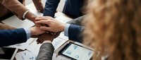 Business team putting hands together in unity gesture over meeting table with financial documents and tablet showing charts and graphs.