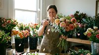 Young woman of South Asian descent, florist wearing olive apron, arranging fresh peach and coral roses in vase surrounded by blooming flowers in studio workspace, natural light from window.