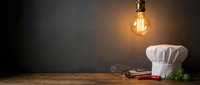 Culinary workspace with chef hat, kitchen utensils and glowing vintage bulb on wooden table against dark background for cooking and restaurant concepts.